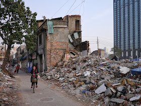 woman riding a bicycle past demolished buildings and a modern tall building in the background