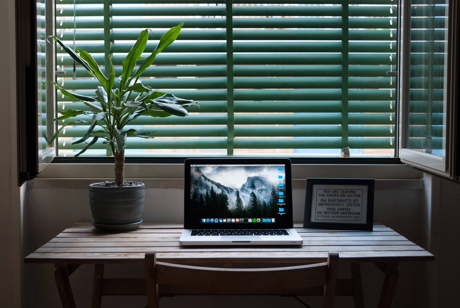 Vista horizontal de uma janela com uma mesa posicionada logo abaixo. Sobre a mesa temos um notebook, um vaso de planta ornamental e um porta-retratos. Sendo que o notebook é o principal equipamento para home office nos dias de hoje.