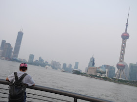 young woman wearing a "BOSS" cap leaning against a railing next to the Huangpu River in Shanghai with the Oriental Pearl Tower in the background
