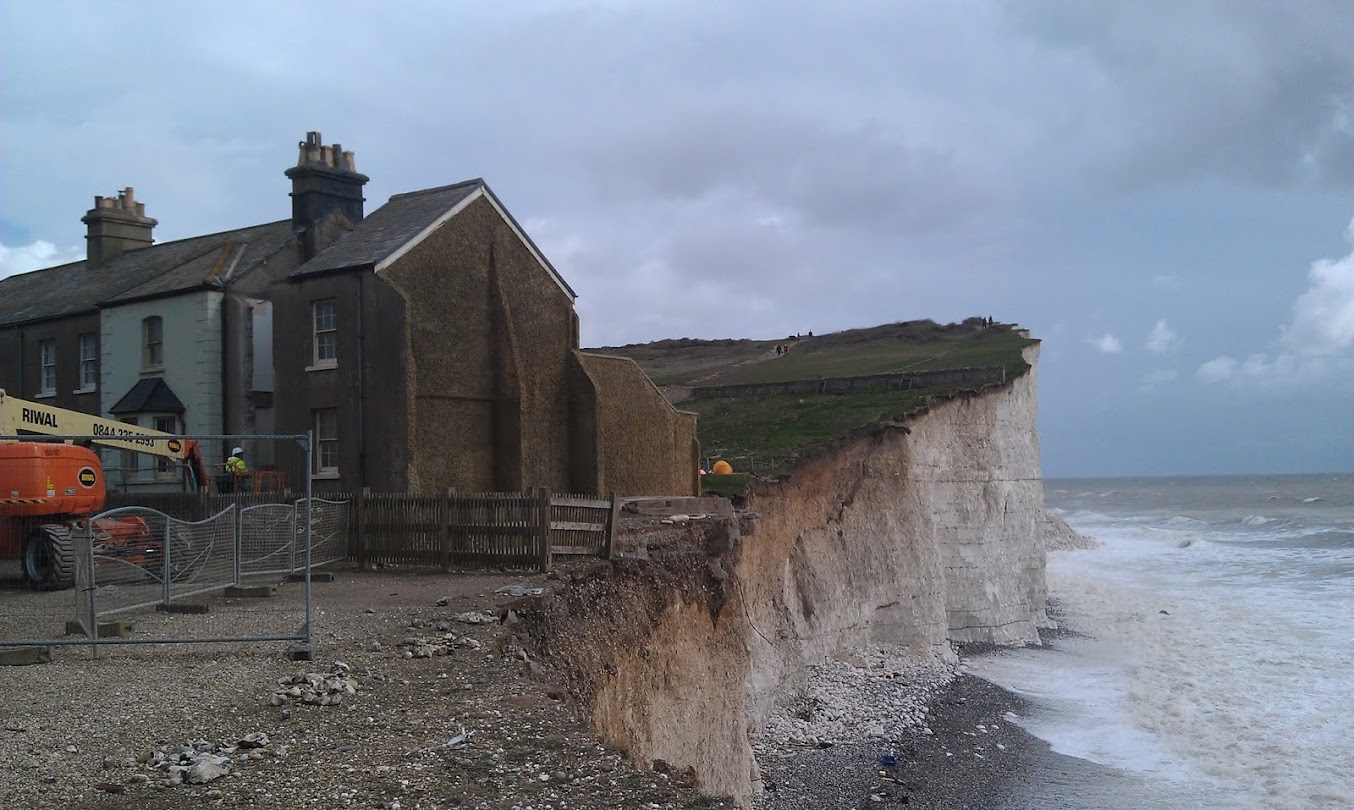 Chalk cliffs at Birling Gap showing coastal erosion.  to the right white capped waves drive against the beach.  To the left are coastguard cottages falling off the cliff.  The end cottage is being demolished before it collapses.