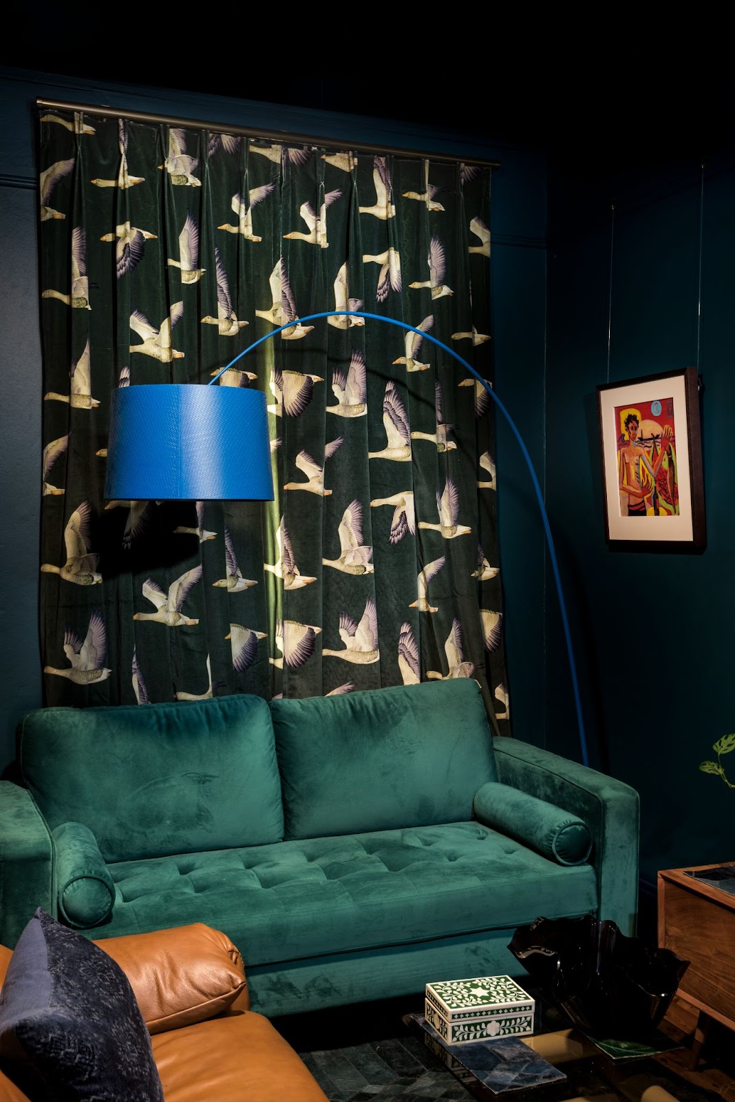 A stylish living room with dark green walls, a teal velvet sofa, a vibrant blue floor lamp, and curtains featuring a bird pattern, complemented by eclectic artwork on the wall.