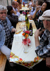 Im Lokal. Tisch mit Gästen, Weinflaschen und -gläser, ein Weizenbierglas.