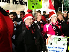 Postlerinnen auf dem Kopf Weihnachtsmamm-Mützen, in den Händen Plakat mit gemaltem Postauto: »Appel am Steuer - Ungeheuer!«.