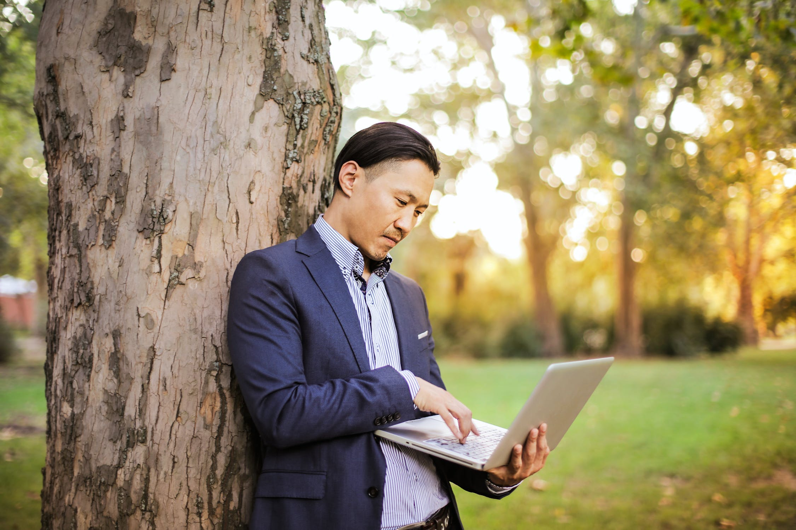 Man in blue suit leaning up against a tree consulting his laptop computer