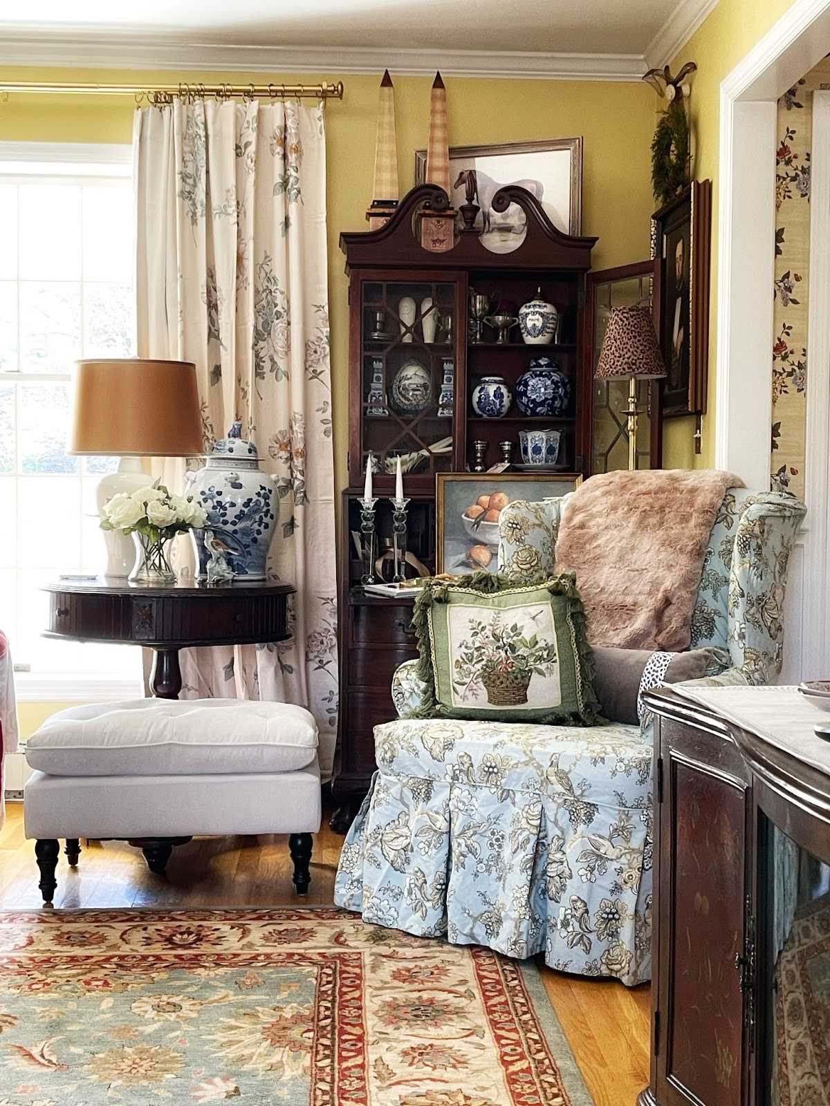 This cozy corner features a beautiful hutch showcasing an array of delicate blue and white porcelain urns and vases, a skirted armchair with a soft floral pattern, a plush ottoman, and small side table.