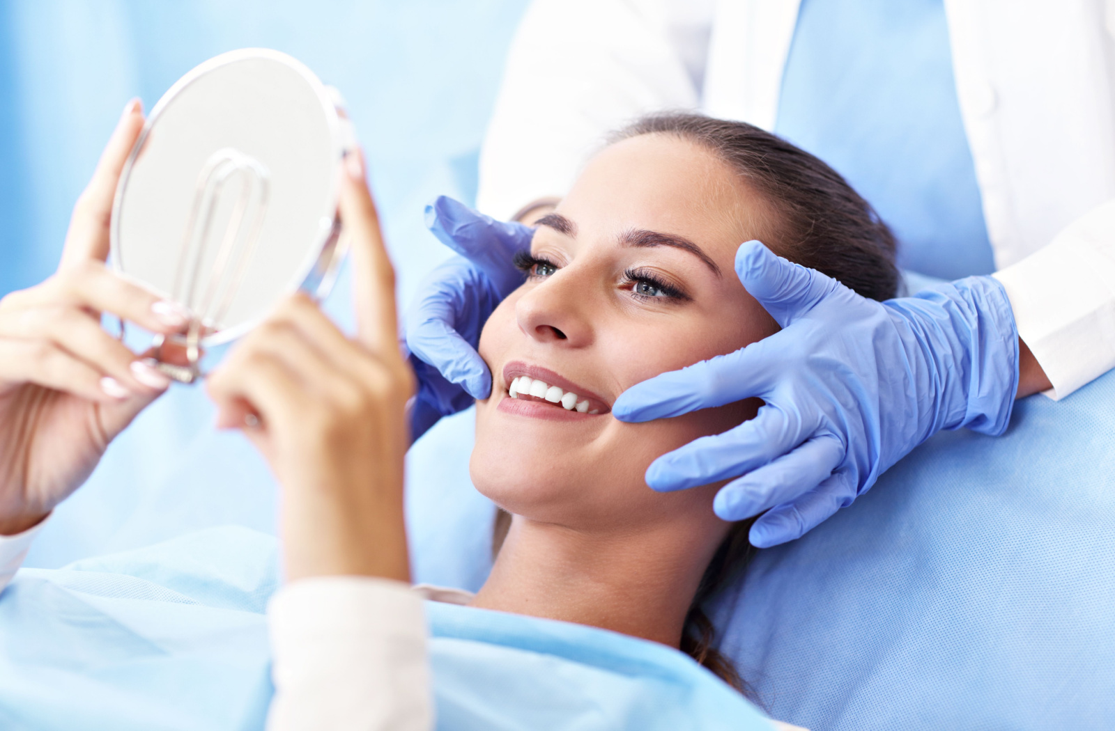 A woman sitting in a dentist’s chair and smiling into a handheld mirror while her dentist gently holds the sides of her face.