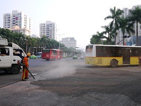 a bus and truck going around a man using a blower to move gravel and creating a large dust cloud on a street in Zhuhai