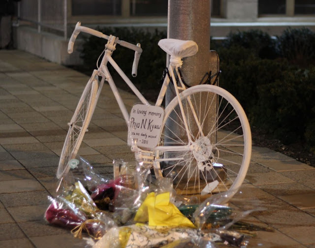 a bicycle painted all white and chained to a lamppost, with a sign on it giving the biker's name and dates, and a pile of flowers and candles in the foreground