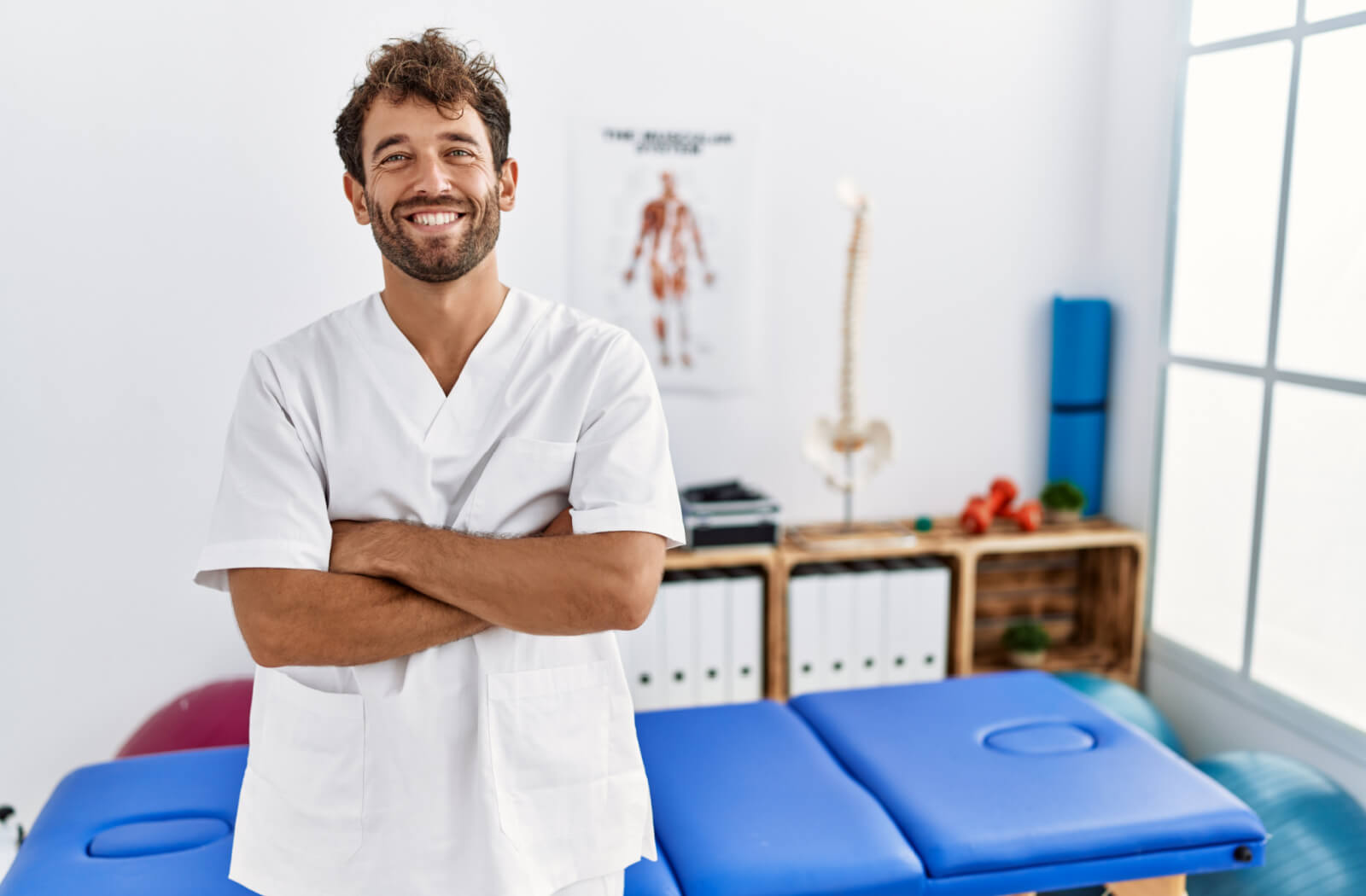 A male physiotherapist in a pain recovery clinic with crossed arms, smiling and looking directly at the camera