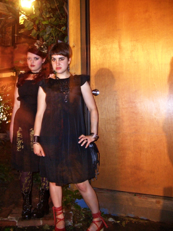 Two girls stand outside against a brown paneled wall. The girl on the left is fat, pale-skinned, and red-haired. The girl on the right is curvy, pale-skinned, and has short, dark hair. They are both in black dresses and black boots.
