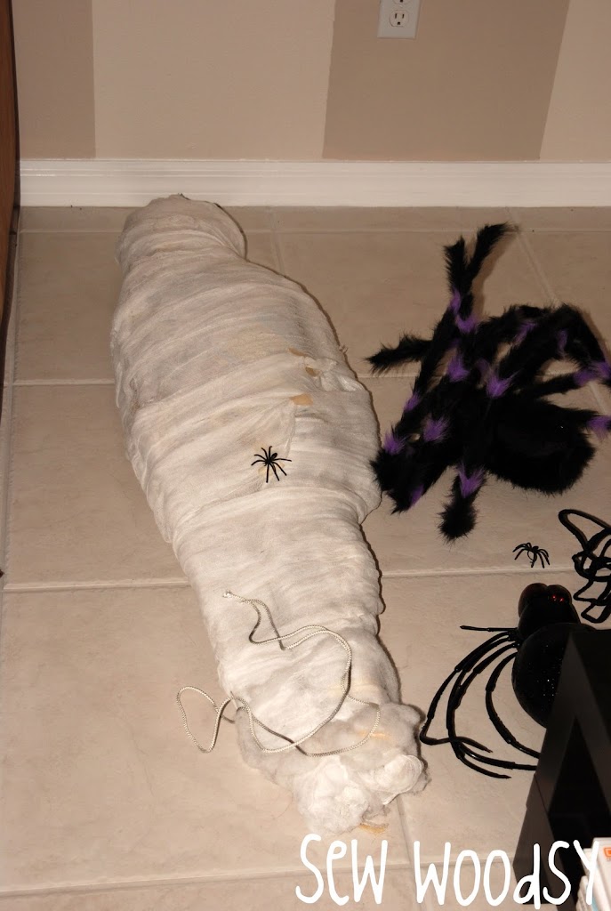 Mummy/webbed person laying on a tile floor with mini spiders on it.