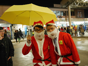 two young men wearing Santa outfits standing under a yellow umbrella