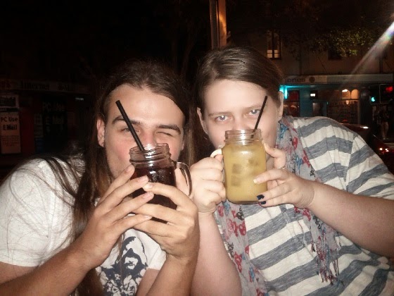 A pale skinned young main with long dark brown hair on the left holds a dark drink in a jamjar up in front of his face.  A pale-skinned young woman on the left holds a yellow drink in a jamjar up to her face.  They are highly amused.