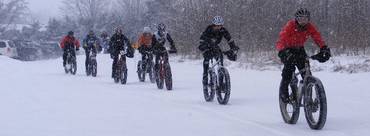 Front, right side view of a winter cyclists, riding Surly fat bikes single filed, in heavy snow beside a bare tree line