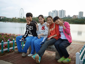 four high school students sitting on a bench with a lake and a ferris wheel in the background in Zhanjiang