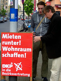 Zwei Genossen befestigen DKP-Plakat an Lichtmast. »Mieten runter! Wohnraum schaffen! DKP in die Bezirksvertretung«.