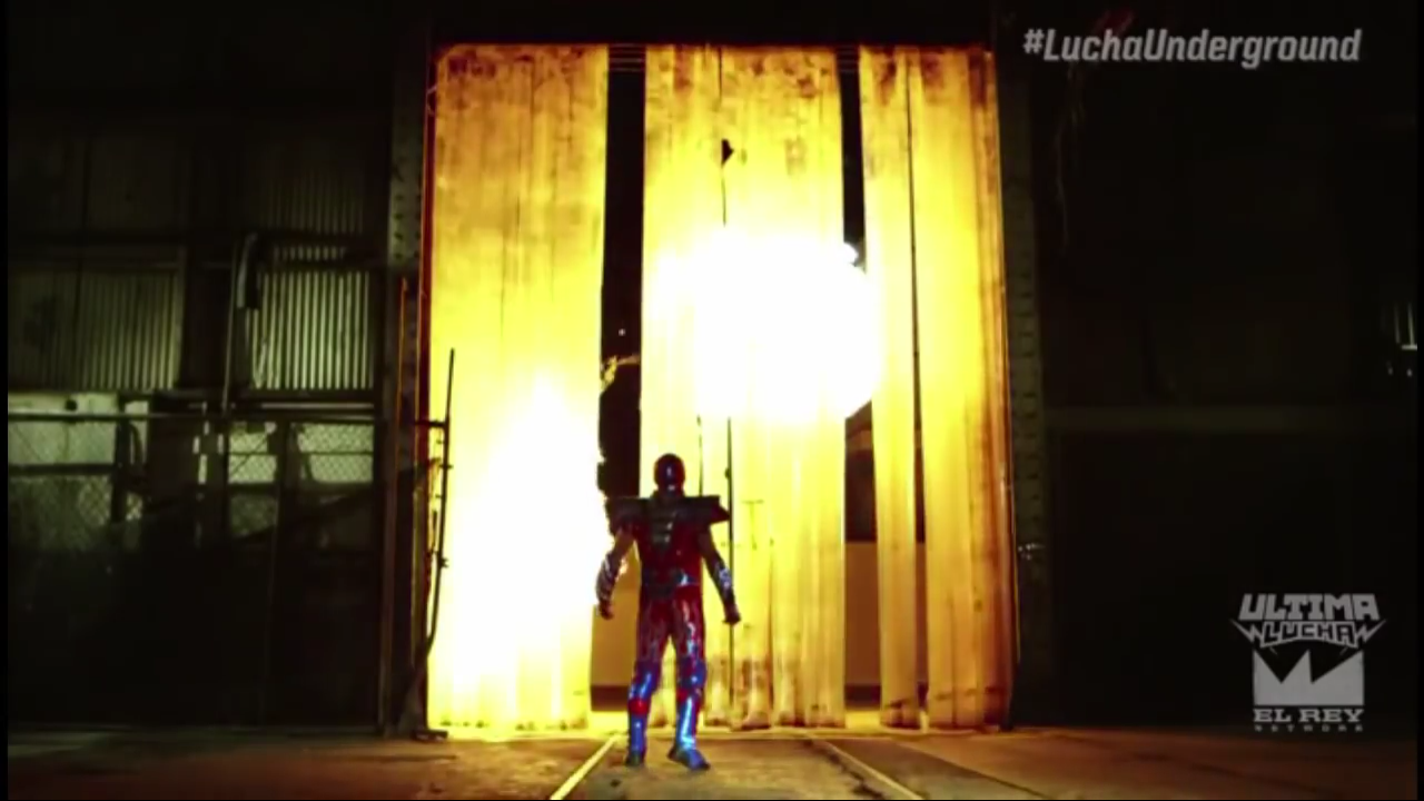 Screen cap from Lucha Underground. Aerostar, a luchador dressed in a metallic red body suit, is in a warehouse and standing in front of large vinyl strips. There is a massive fireball behind the strips; the fireball is intended to represent Drago, a humanoid dragon luchador, taking off for parts unknown.