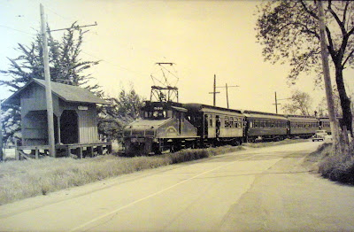 1824 - P&SR train at the Bassett Station (close to Fredericks Road) south of Sebastopol. Engine 506 is pulling four passenger cars with many people hanging out the windows of the cars and a line of 1930's autos on the road next to the train. Since the P&SR ended passenger service in the early 1930's, this may have been one of the final runs carrying passengers.