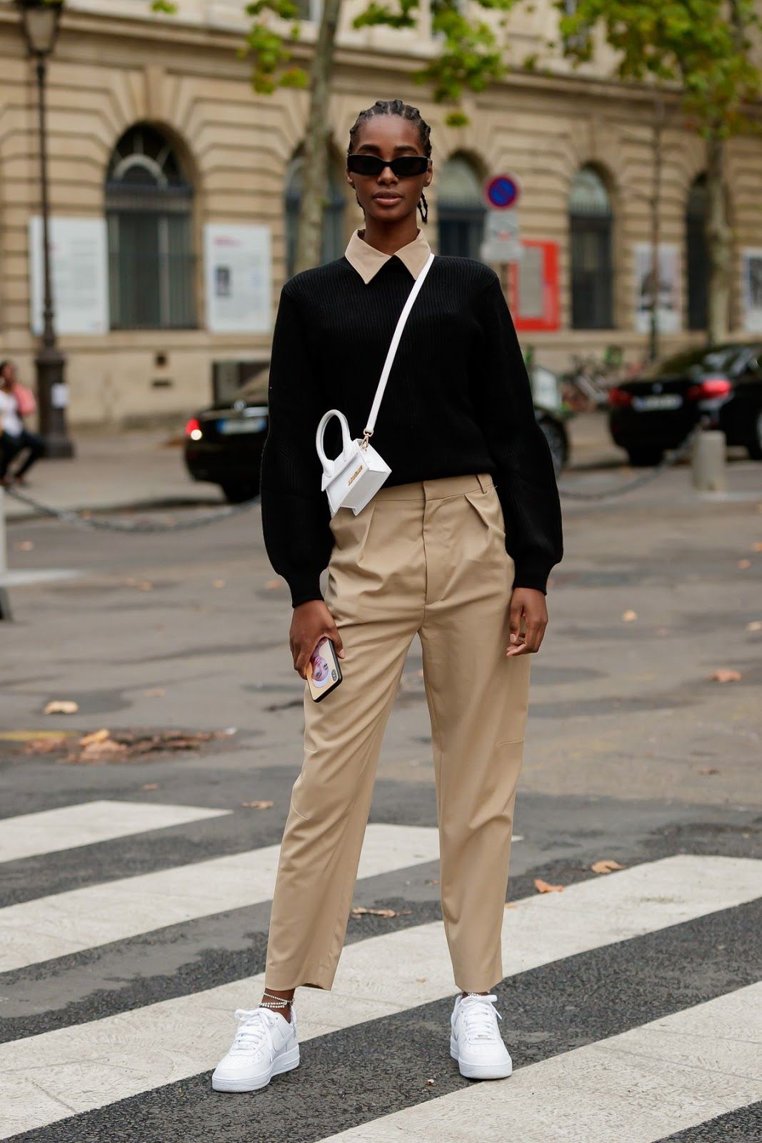 Modelo negra, de cabelos pretos trançados. Ela está em um cenário urbano, utilizando uma camiseta de uma calça cargo bege. Por cima, ela usa um suéter e uma mini bag branca. 