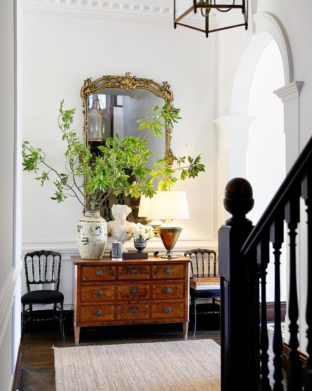 A bright and elegant hallway with a wooden dresser topped with decorative items, a large ornate mirror, and two black chairs, all illuminated by natural light from a nearby window