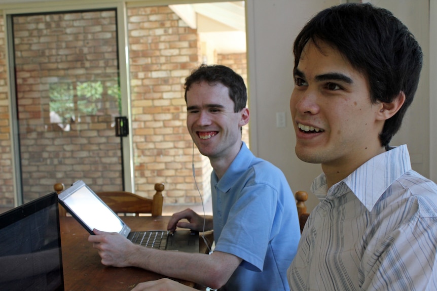 Two men sit at a wooden kitchen table. One is looking to distance while the other has his hands on an open laptop
