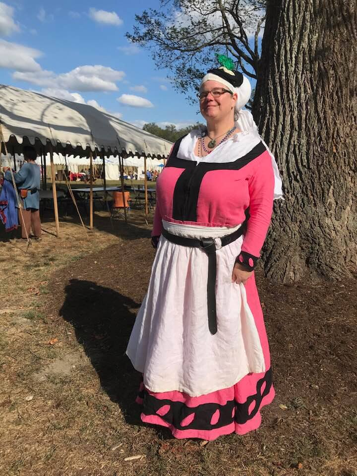 [A woman stands in three quarters profile with her back to a large brown tree trunk. She is wearing a pink and black dress, with a white apron, a white undershirt, and an illustrious white veil with black hat. She is smiling at the camera.]
