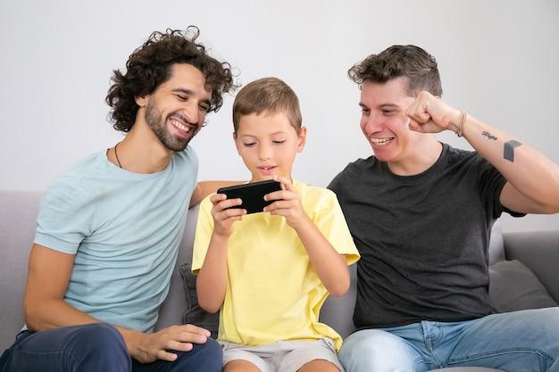Free Photo | Excited boy playing game on mobile phone, his two happy dads  sitting near him and helping. front view. family at home and communication  concept
