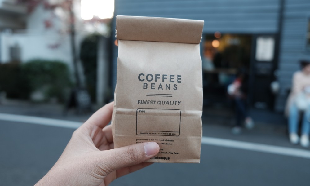 Unbleached kraft paper coffee bag filled with roasted coffee beans with black text and coffee shop in background.  