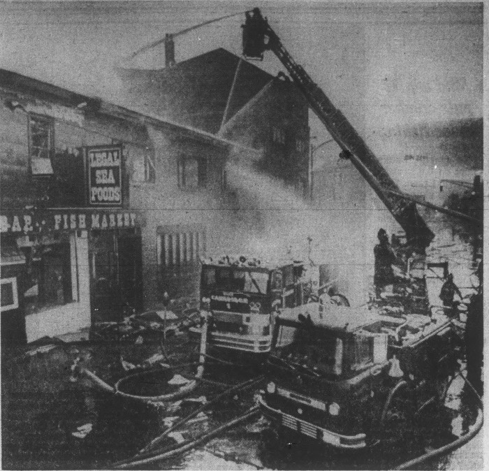Black and white newspaper photo of two fire engines with hoses out. One had a ladder spraying onw a two story building. Awning on building reads "Fish market" and sign reads "Legal Sea Foods."