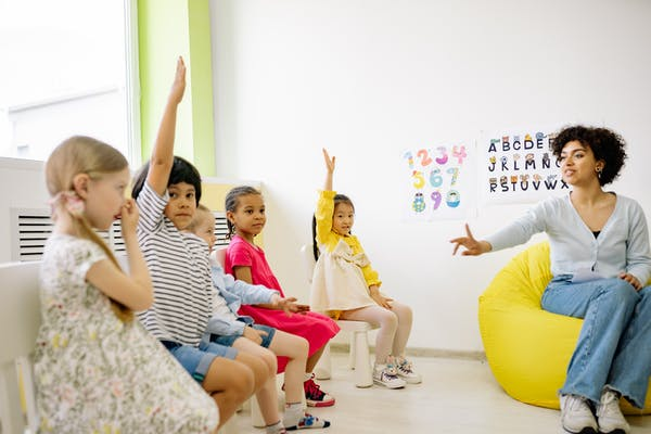 Children raising their hands and the teacher pointing to a child to respond. 