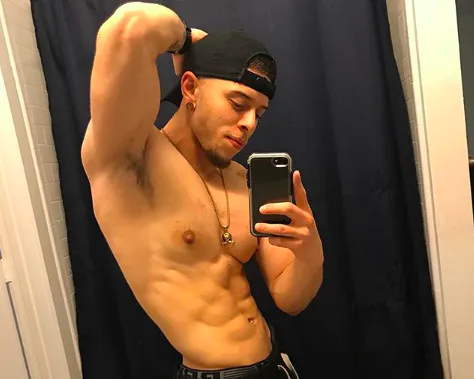 A shirtless young man with tan skin, a small chin beard, a backwards baseball cap, and a pendant on a chain hits a classic thirst trap pose with one hand behind his head and his shirtless body slightly in profile, showing off his perfectly defined muscles. He holds his camera in the other hand to take this mirror selfie, and looks down at the screen, smiling slightly, giving the impression that he knows exactly what a thirst trap he is.