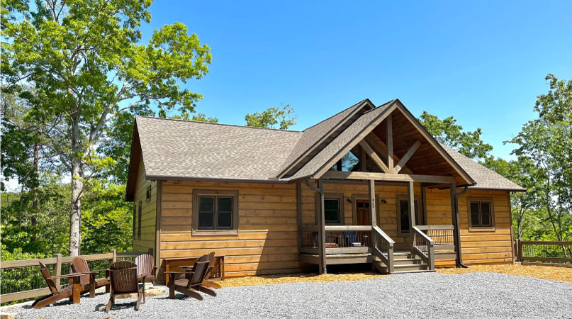 A wood cabin-style home in Ellijay, GA, on the side of a mountain. There are wooden Adirondack chairs surrounding a fire pit outside and the driveway is filled with gravel.