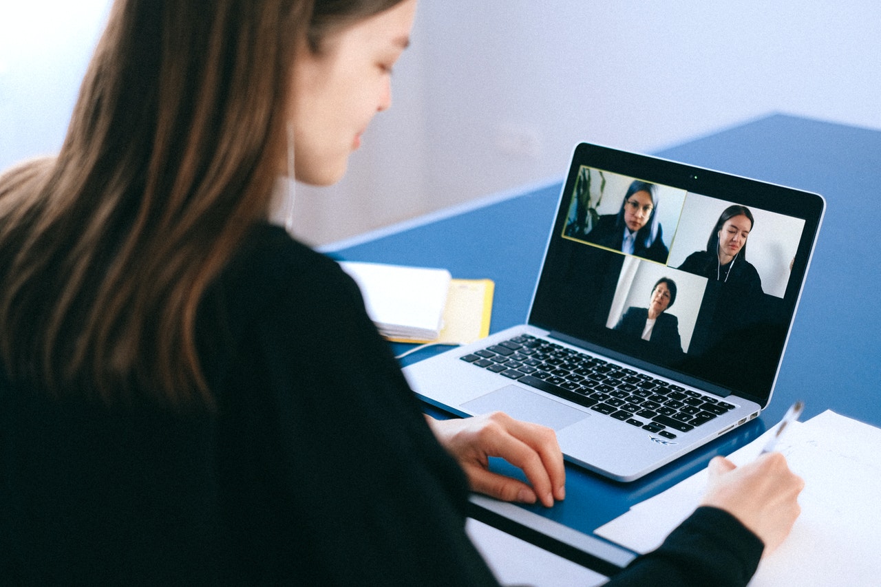 A imagem mostra uma mulher branca, de cabelo castanho-claro liso na altura dos ombros, em frente a um laptop prata. Na tela, é possível ver uma chamada de vídeo, na qual há mais três mulheres. 