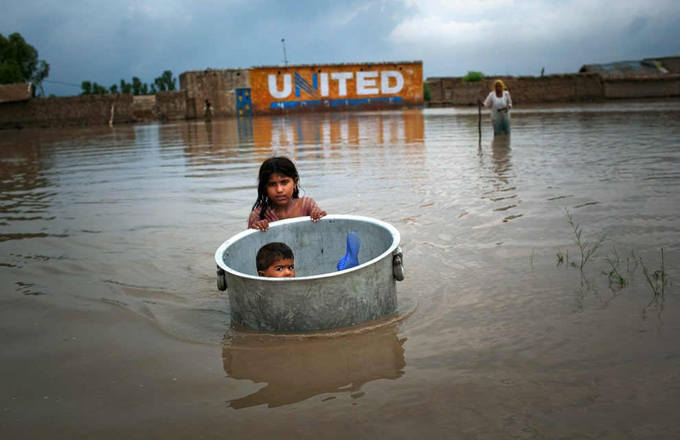 A young Pakistani girl wades through floodwater pushing a giant metal pot with a smaller boy inside.