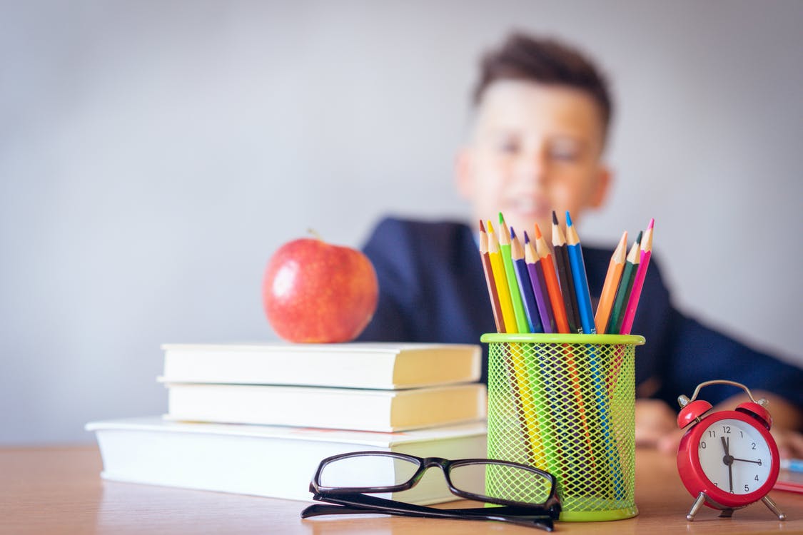 A close up of a school desk with books, glasses, an apple and colour pencils on it, with an out of focus boy in the background