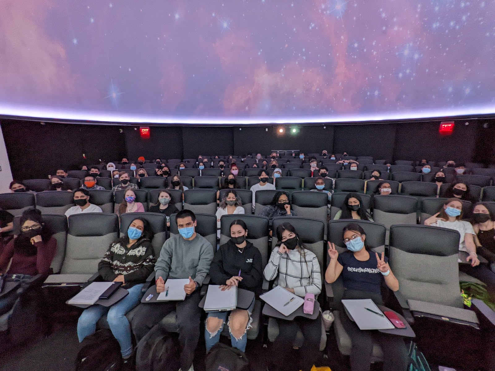 Photograph of the Sac State planetarium, with around two-thirds of the seats occupied by excited students wearing masks and taking notes. They are sitting under the planetarium dome which looks to be projecting an image of a nebula or something similar.