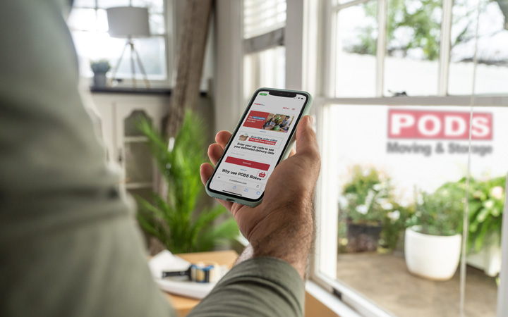 Close up of a man holding his cell phone and viewing the home screen of PODSboxes.com. He is about to order moving supplies and have them delivered right to his house. There is a PODS moving and storage container in the driveway which is visible through the window.