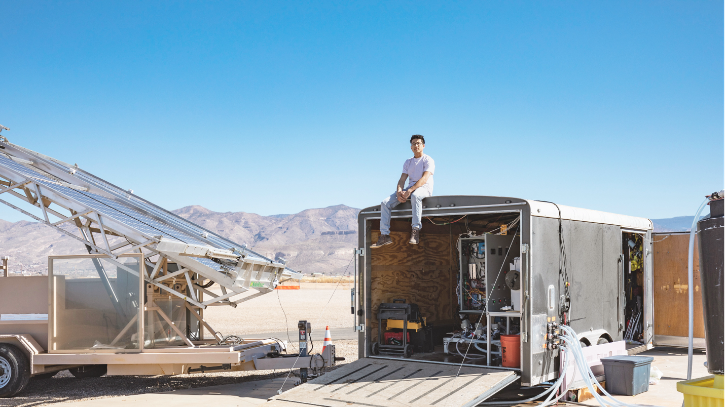 Jonathan sits on the roof of an open trailer, surrounded by cords and pipes. In the background, mountains rise under a light blue sky.