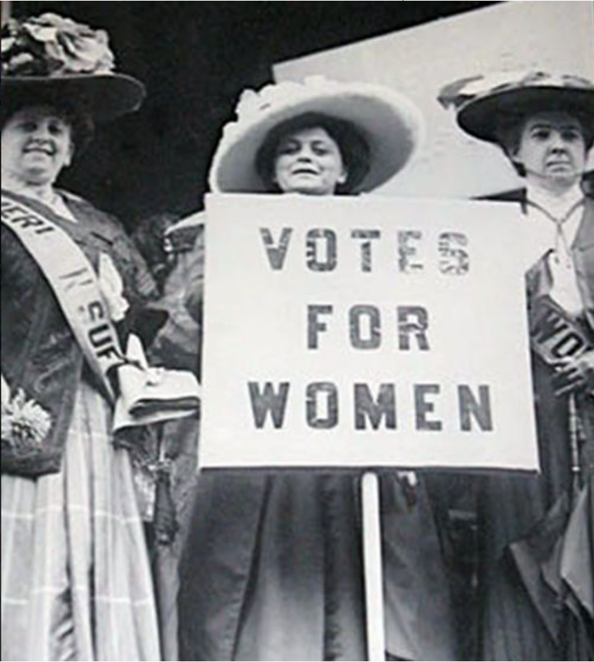 The text on the sign reads “Votes for Women”. The three women wear long dresses and wide brimmed hats with elaborate decorations on top. The women to the left and right wear sashes with illegible text (only some letters are readable: SUF; WO); the woman in the centre stands behind the large sign.