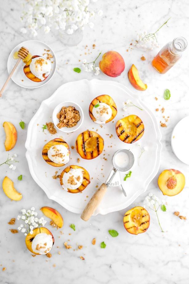 overhead shot of five grilled peaches on white platter with more grilled peaches around, peach slices, and mint leaves