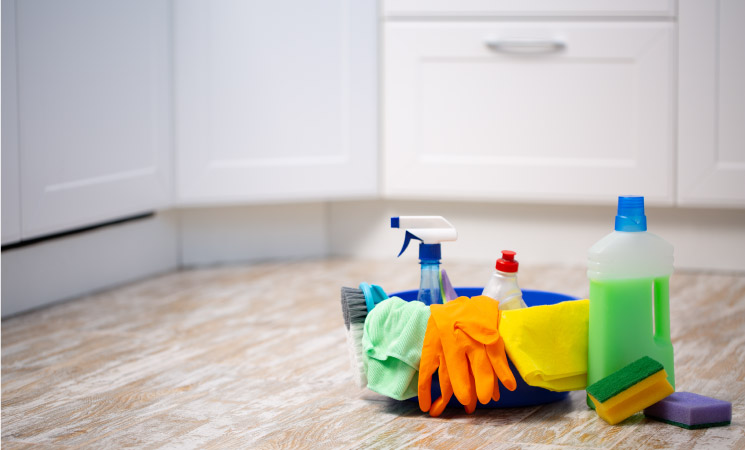 A large blue bowl on the floor of a clean kitchen. The bowl has various cleaning supplies in and around it, including soap, window cleaner, a scrub brush, rags, rubber gloves, another cleaning solution, and two colorful sponges. 