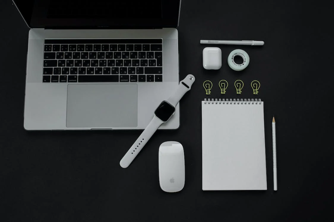 A smartwatch, a laptop, a mouse, airpods, and a notepad with a pencil
