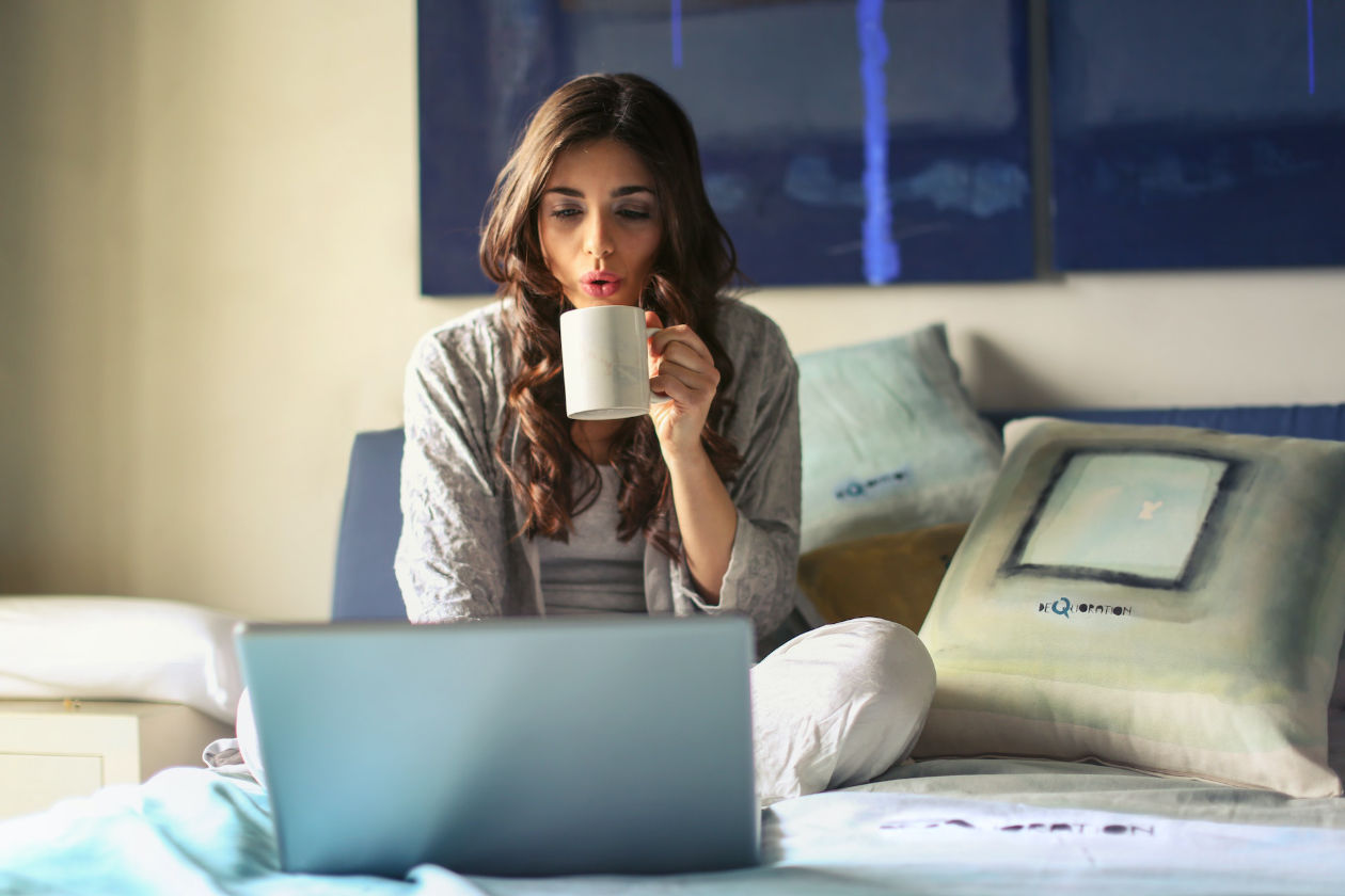 Woman with mug sitting on bed while looking at laptop