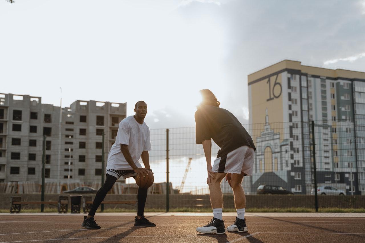 La imagen muestra a dos hombres en una cancha de baloncesto al aire libre. De izquierda a derecha, hay un hombre negro mirando a la cámara y sosteniendo la pelota de baloncesto. Frente a él y de espaldas a nosotros se encuentra un hombre blanco, con el pelo hasta la nuca, que está ligeramente encorvado y con las manos en las rodillas.