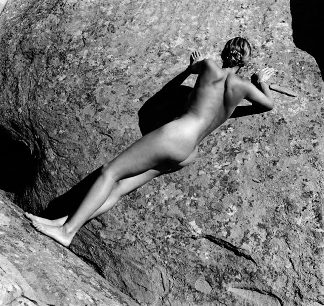 A fotografia em preto e branco é composta por uma mulher branca, nua, de costas e com os cabelos presos num coque baixo, apoiada numa grande pedra que ocupa todo o fundo da foto.