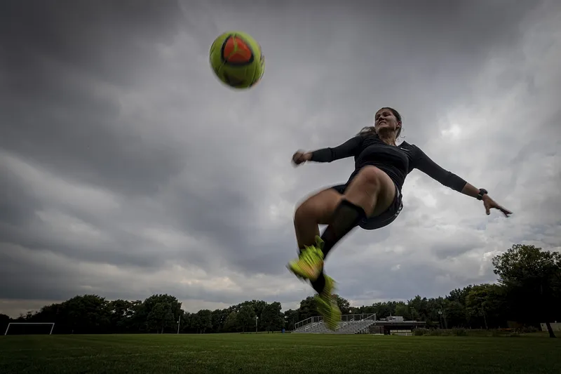 Imagem de uma mulher, num campo de futebol, vestindo roupa de ginástica preta. Ela está no ar, durante o movimento de chutar uma bola