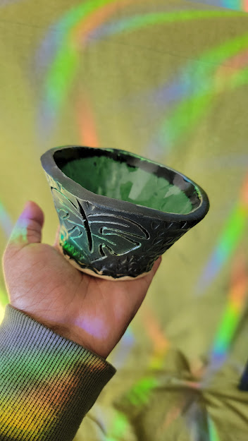 Photo of my left hand holding up a small piece of pottery made to hold a small plant. The pottery is green on the inside and black on the outside and has a monstera leaf shape carved into the side.