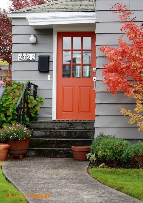 Fachada de casa com porta laranja, paredes cinza e jardim.