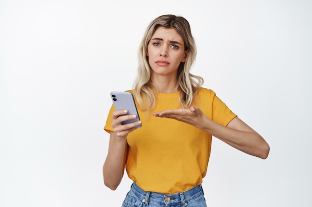 Free photo troubled young woman asking help with mobile phone pointing at smartphone and looking upset standing over white background