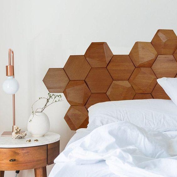 Quarto com cama de casal com cabeceira de revestimento hexagonal amadeirado, parede fundo branca, mesinha de canto amadeirado com bancada branca e abajur.